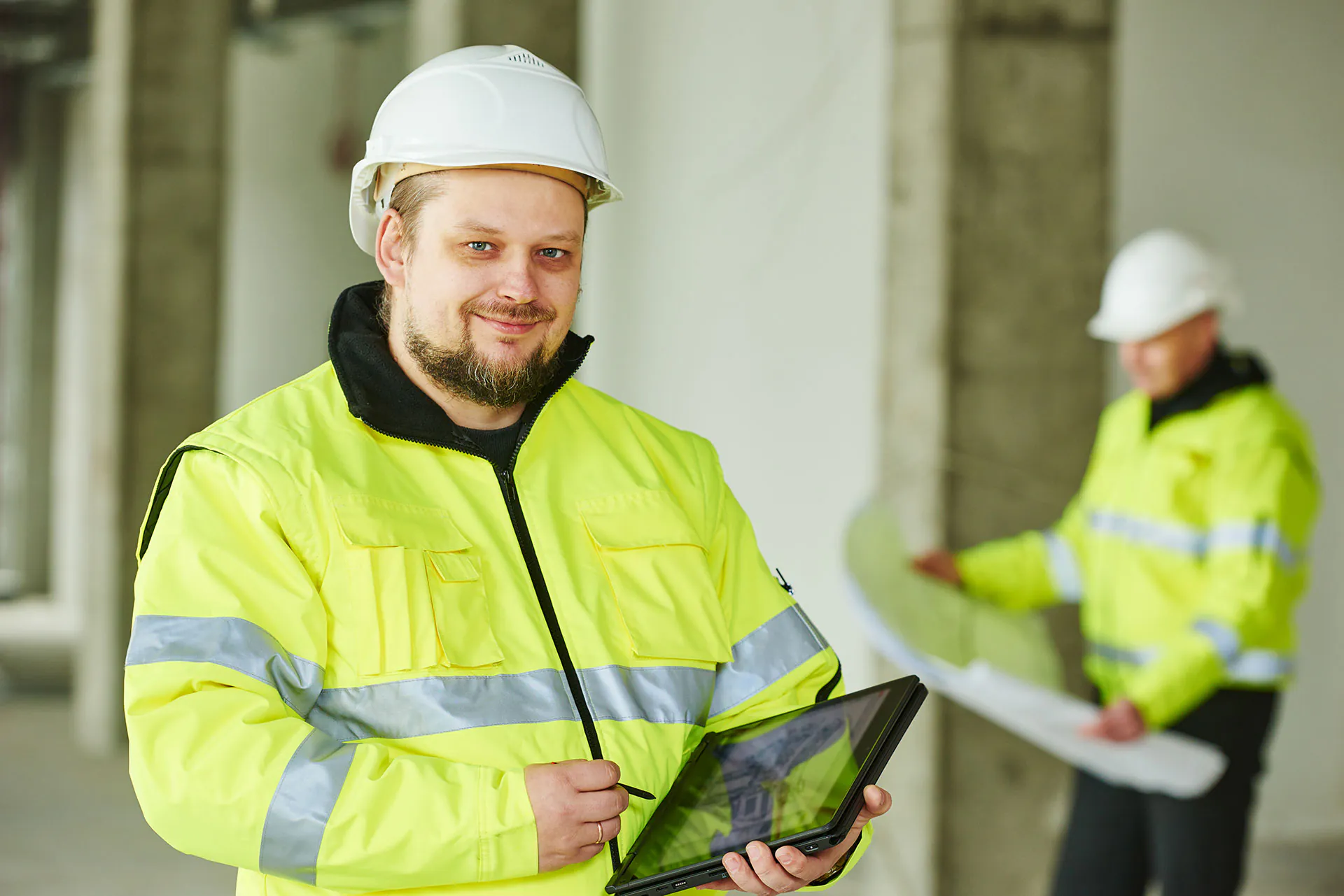 Een technicus in een reflecterend geel veiligheidsjack houdt een tablet vast voor field service management terwijl hij in een bouwproject met betonnen zuilen poseert.