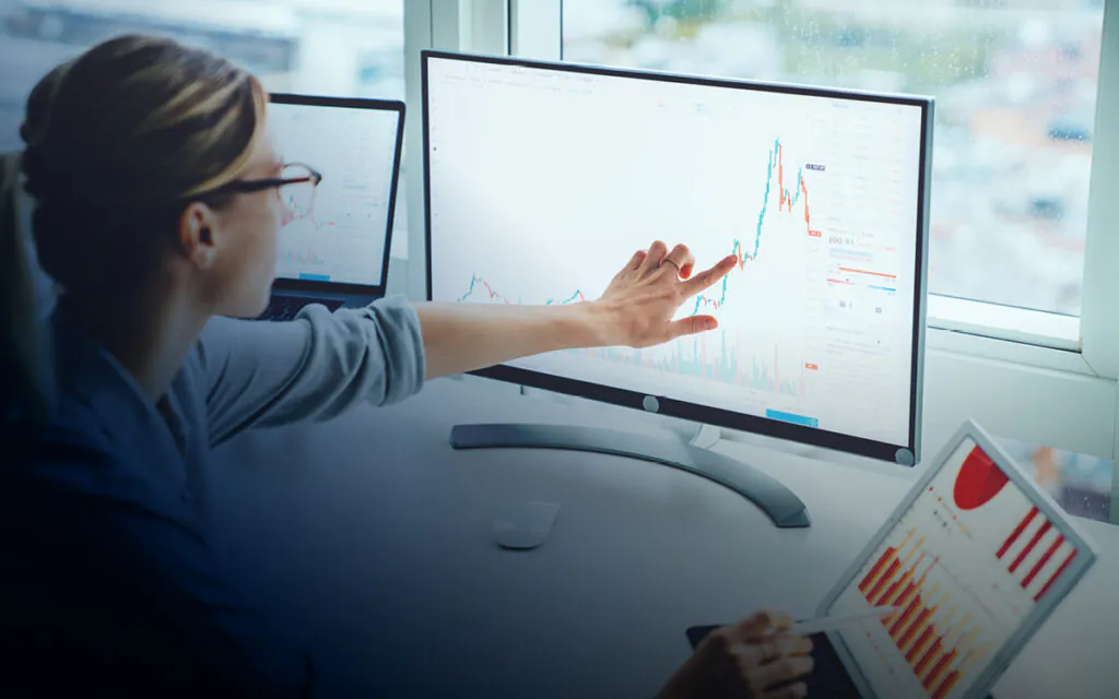 Een analist wijst naar een grafiek op een beeldscherm ter ondersteuning van **asset management**, zittend voor een raam met meerdere apparaten die financiële gegevens tonen.