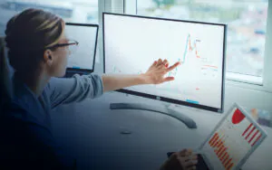 Een analist wijst naar een grafiek op een beeldscherm ter ondersteuning van **asset management**, zittend voor een raam met meerdere apparaten die financiële gegevens tonen.