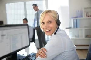 Een glimlachende blondine met een headset kijkt achterom terwijl zij werkt op een computer in een kantoor, wat haar ondersteunt bij taken gerelateerd aan SAP FSM en effectief field service management.