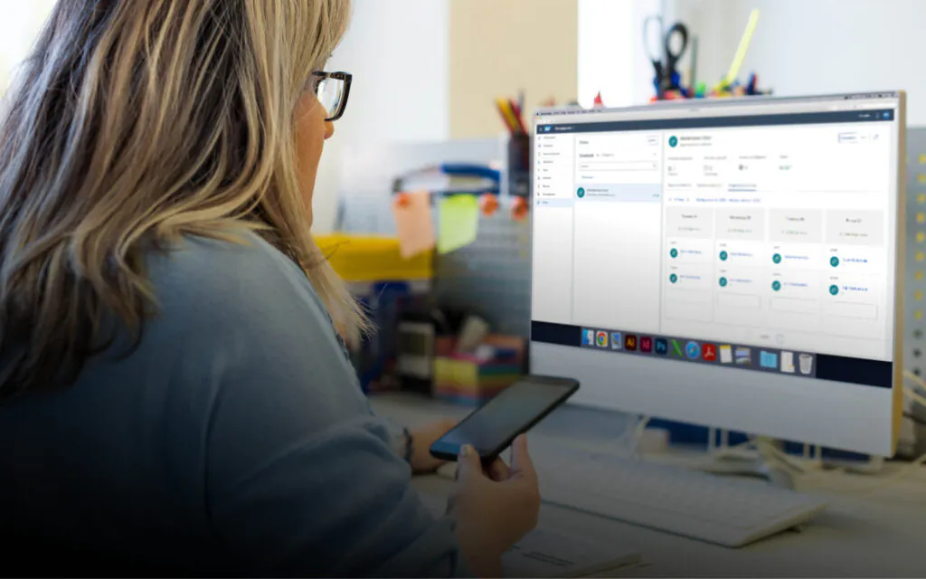 Een vrouw met een bril bekijkt SAP FSM-software op een computerscherm en gebruikt haar smartphone in een kantooromgeving voor efficiënt asset management en dagelijkse werkzaamheden.