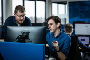 Twee collega's bespreken SAP FSM-processen op een computerscherm in een modern kantoor, waarbij de een naar de monitor wijst en de ander aandachtig toekijkt. Tekst: PHILIPS.