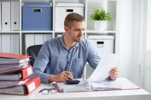 Een man controleert documenten en gebruikt een rekenmachine voor efficiënt asset management aan een bureau in een kantoor vol met ordners en opbergdozen op de achtergrond.