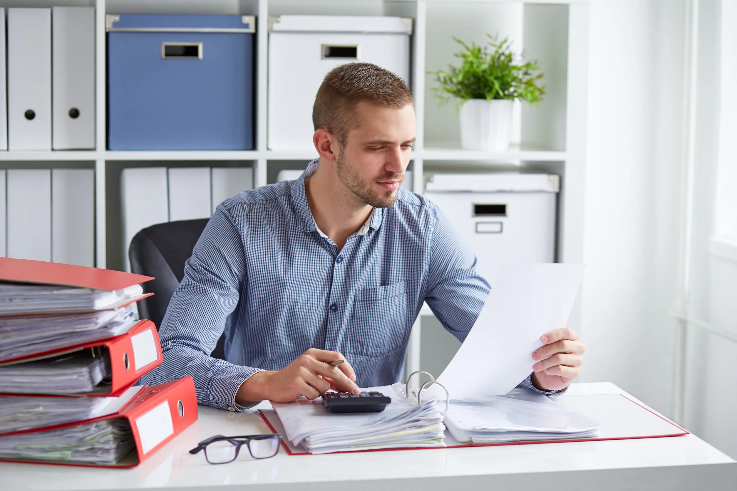 Een man controleert documenten en gebruikt een rekenmachine voor efficiënt asset management aan een bureau in een kantoor vol met ordners en opbergdozen op de achtergrond.