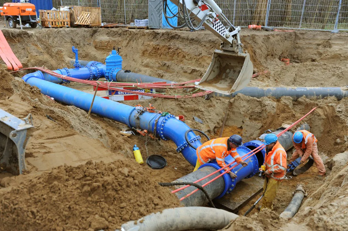 Drie arbeiders in werkkleding installeren blauwe leidingen in een open bouwput onder toezicht van een graafmachine, een essentieel proces voor effectief asset management op de bouwplaats. Tekst op het hekwerk: "HUURHANS