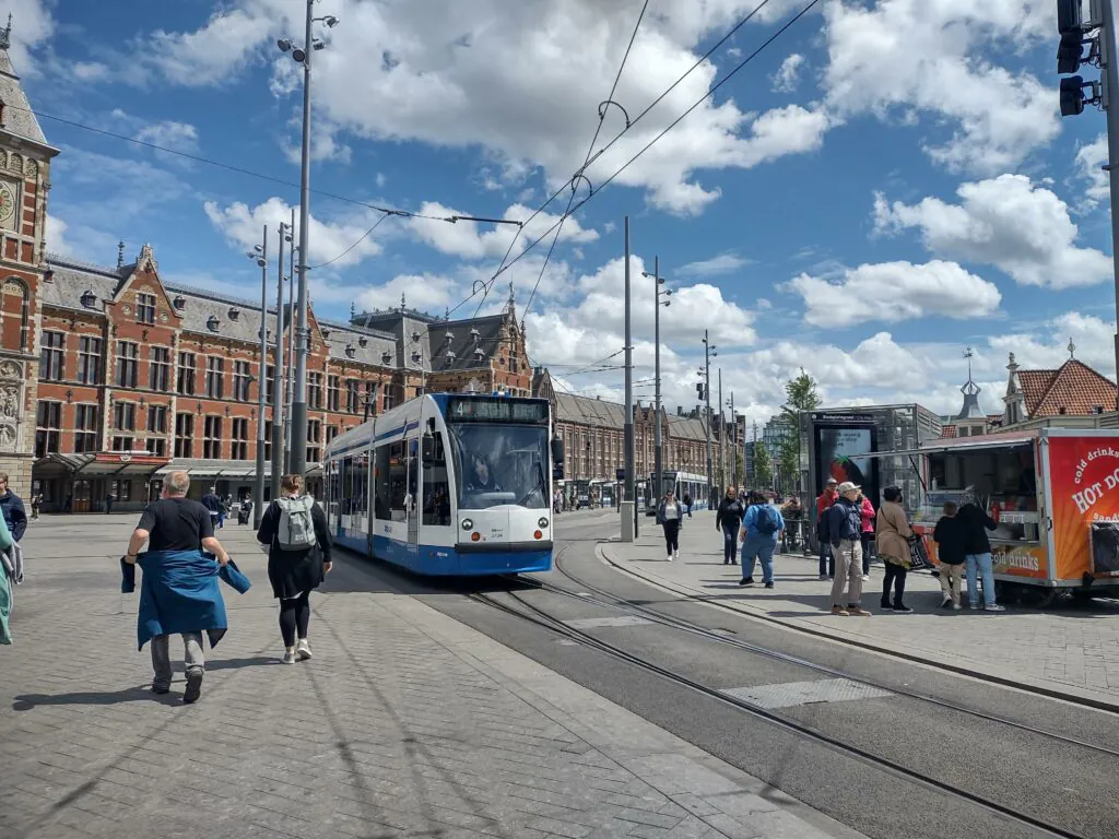 Een blauw-witte tram rijdt over rails langs voorbijgangers en een eetkraam op een druk Amsterdams stationsplein, wat efficiënt asset management in een stedelijke omgeving onderstreept. Tekst op kraam: HOT DOGS.