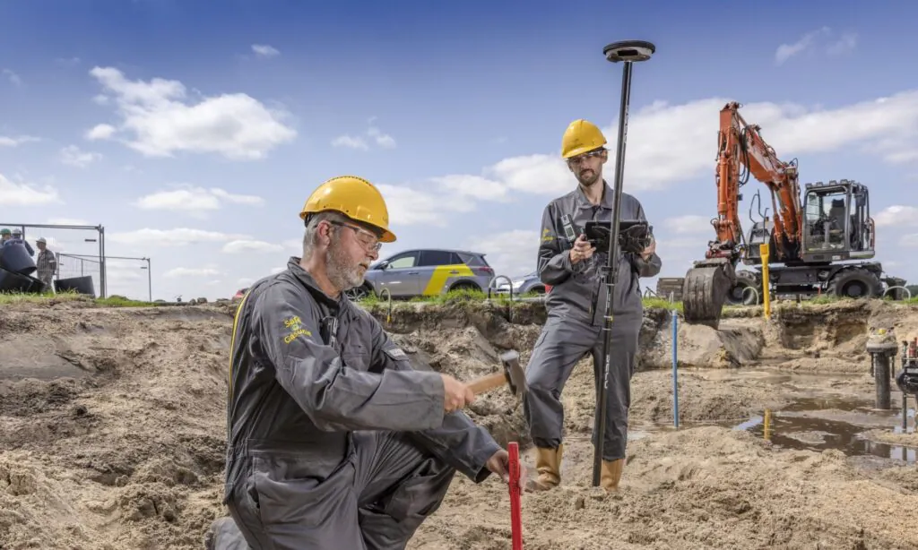 Twee technici in beschermende kleding voeren veldwerk uit aan een pijpleiding met behulp van meetapparatuur en een hamer. Dit ondersteunt efficiënt field service management op een actieve bouwplaats. Tekst op de kleding: "Safe @ Gasunie