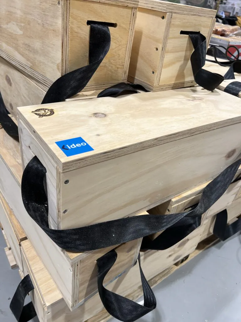 Houten kisten met zwarte draagbanden liggen gestapeld voor efficiënt asset management, waarbij het "ideo" logo op een blauwe sticker op de voorste kist prijkt.