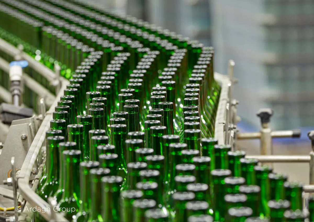 Groene glazen flessen bewegen in een strakke formatie over een lopende band, wat de efficiëntie in asset management binnen een industriële productieomgeving benadrukt. Tekst in afbeelding: © Ardagh Group