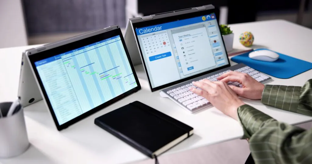 Iemand typt op een toetsenbord bij twee laptops die SAP FSM-planningen en kalenders tonen op een wit bureau in een kantooromgeving. **Tekst in de afbeelding:** Calendar, Oct, 5, 6, 7, 1, 2, 3, 4, 8, 9, 10, 11, 12, 13, 14, 15, 16, 17, 18, 19, 20, 21, 22, 23, 24, 25, 26, 27, 28, 29, 30, 31, Create Task, Team Meeting, Add.