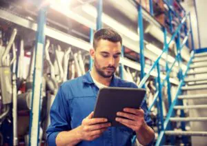 Een monteur in blauwe werkkleding bekijkt een tablet voor efficiënt field service management terwijl hij in een magazijn vol met metalen onderdelen op stellingen staat.