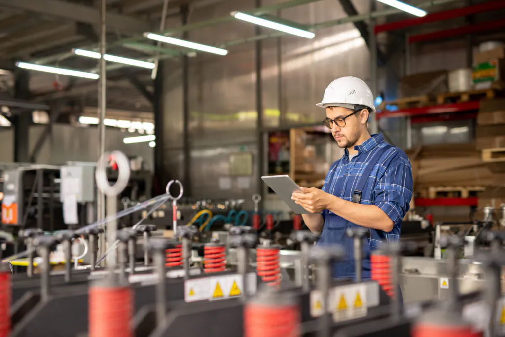 Een technicus met een helm bekijkt een tablet voor field service management terwijl hij naast industriële machines in een magazijn staat, omringd door stellages en opslagmaterialen.