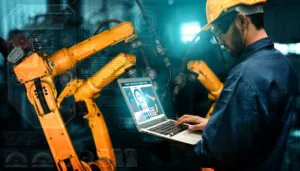 Een technicus in werkkleding en helm bedient een laptop met SAP FSM-software voor efficiënt asset management, terwijl hij toezicht houdt op gele robotarmen in een geautomatiseerde industriële omgeving. **Tekst op scherm:** SMART INDUSTRIAL CONTROL PANEL Performance 40% 25% 90%
