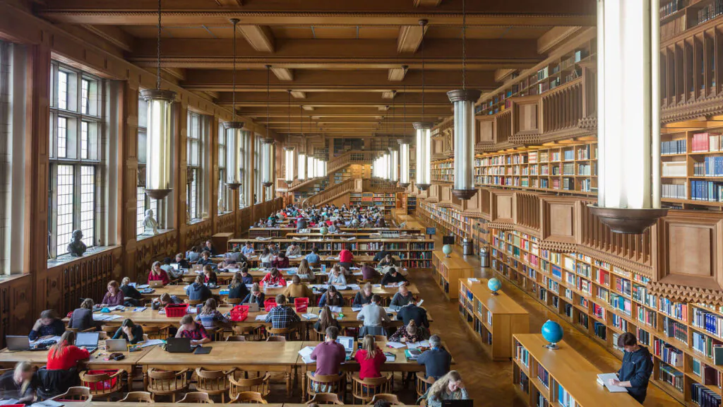 Studenten studeren aan lange houten tafels in een uitgestrekte, klassieke bibliotheek met torenhoge boekenplanken, wat een stille omgeving creëert die ideaal is voor concentratie en efficiënt asset management van informatie.