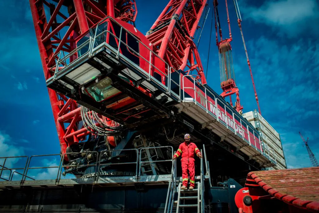 Een technicus in rode kleding daalt de trap af van een enorme rode Mammoet-kraan voor asset management onder een blauwe lucht. Tekst op de kraan: "MAMMUT", "LIEBHERR", "A