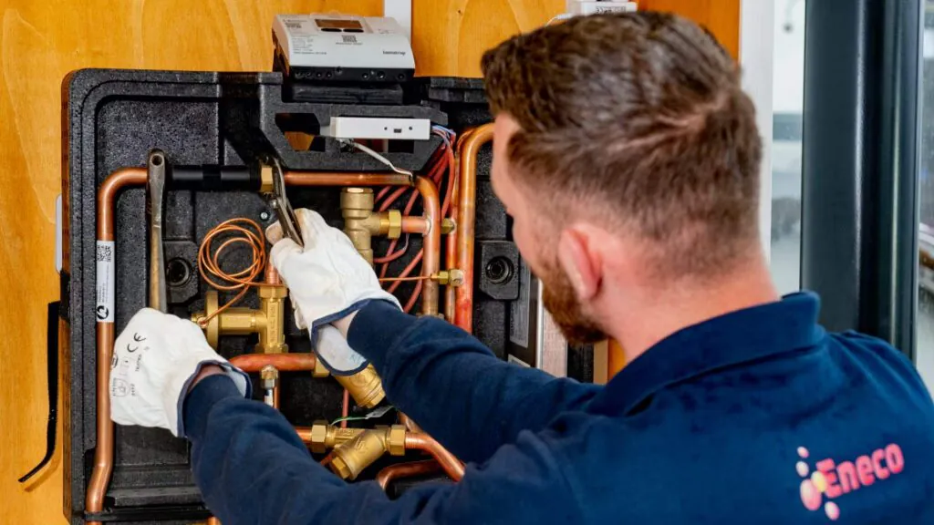 Een technicus met werkhandschoenen voert onderhoud uit aan een warmtepompinstallatie met koperen leidingen en sensoren, wat essentieel is voor effectief field service management. Tekst in de afbeelding: "kamstrup", "CE", "Eneco".