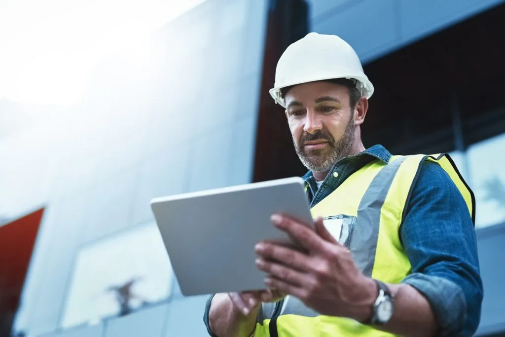 Een bouwvakker met veiligheidshelm gebruikt een tablet voor efficiënt field service management voor een modern kantoorpand, terwijl hij zorgvuldig naar het scherm kijkt bij daglicht.