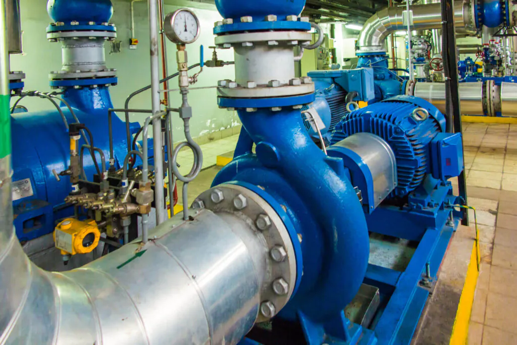 Blauwe industriële pompen en elektromotoren functioneren in een machinekamer, wat essentieel is voor effectief asset management binnen een industriële installatie met metalen leidingen en meetinstrumenten. Tekst: PO 1