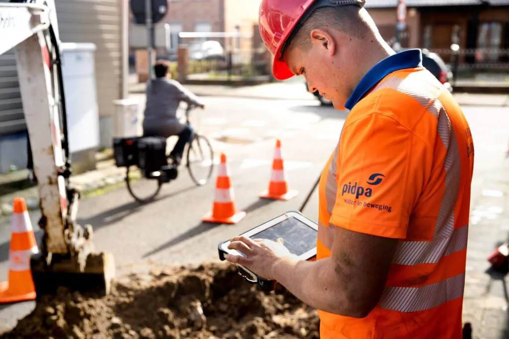 Een technicus met helm en reflecterend oranje uniform gebruikt een tablet voor field service management nabij een bouwput, terwijl een fietser op de achtergrond voorbijrijdt. Tekst op shirt: "pidpa water in beweging".