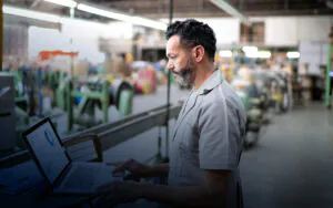 Een technicus in werkkleding bekijkt een laptop met financiële rapportages voor asset management terwijl hij in een industriële fabriekshal staat. Tekst op scherm: Financial Projection Report.