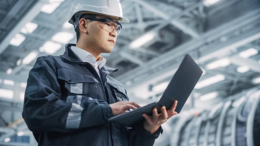 Een technicus met helm en veiligheidsbril werkt op een laptop voor efficiënt field service management in een moderne, industriële fabriekshal vol met complexe machines en apparatuur.
