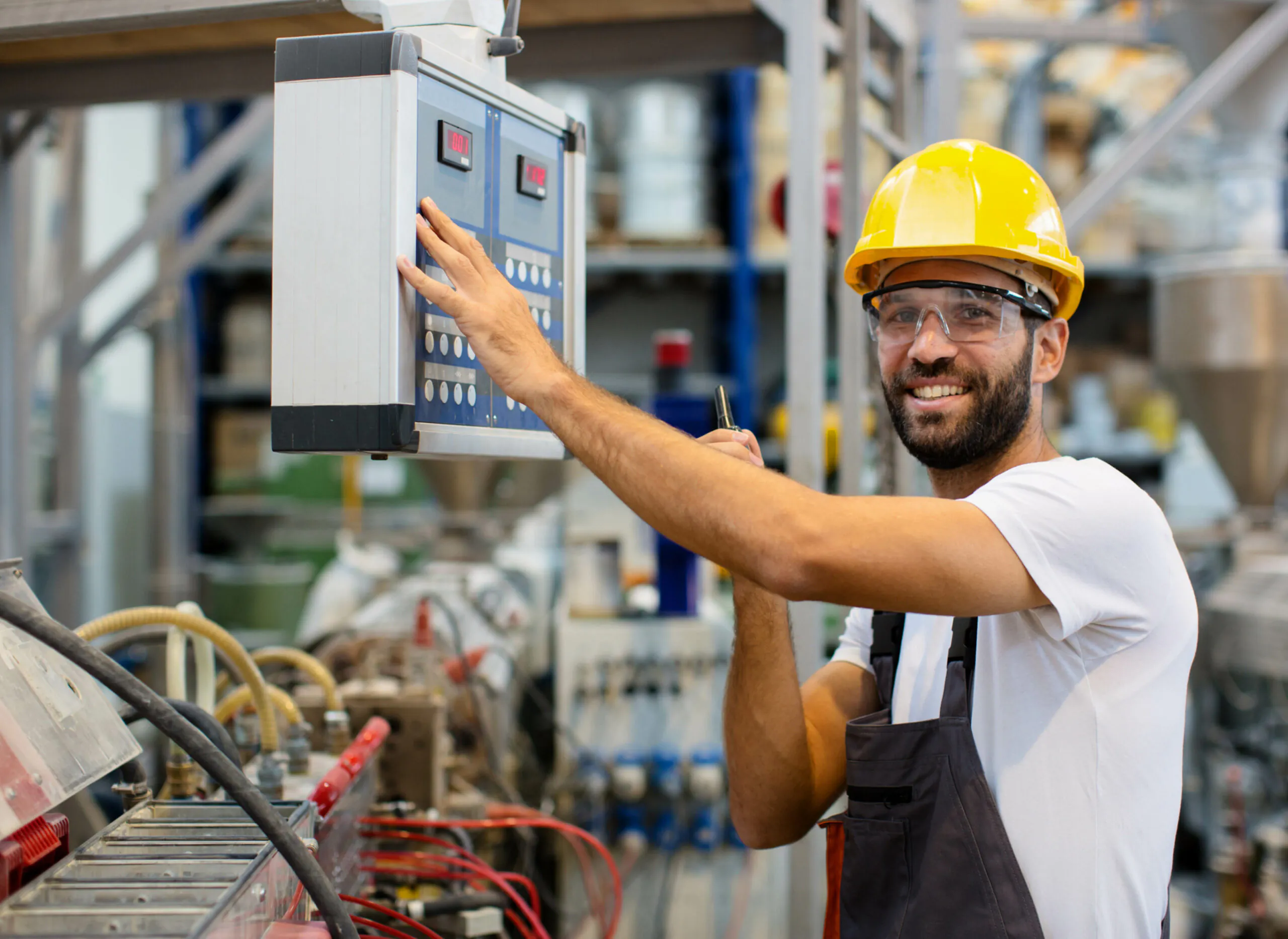 Een glimlachende technicus met helm en veiligheidsbril bedient een bedieningspaneel in een magazijn, wat bijdraagt aan efficiënt field service management bij het onderhoud van industriële machines.