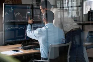 Twee collega's analyseren aandelenkoersen op meerdere monitoren; de zittende persoon wijst naar een grafiek, wat helpt bij strategisch asset management in een modern kantoor achter een glazen wand.