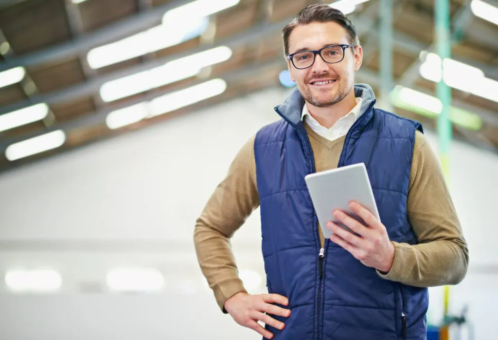Een glimlachende man met een bril houdt een tablet vast voor field service management terwijl hij in een ruime, verlichte industriële hal staat.