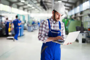 Een technicus met helm typt op een laptop voor field service management, terwijl collega's in de achtergrond van een industriële fabriekshal aan het werk zijn.