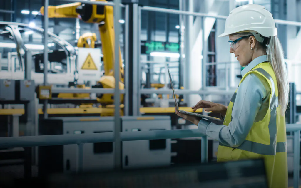 Een ingenieur met een veiligheidshelm bedient een laptop voor **field service management** op een geautomatiseerde productielijn met gele robotarmen en waarschuwingsborden met de tekst "DANGER".