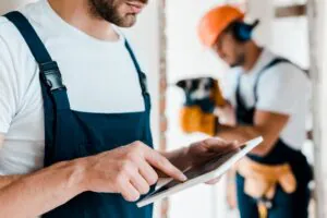 Een bouwvakker in werkkleding bedient een tablet voor SAP FSM, terwijl op de achtergrond een collega met een boormachine aan het werk is op een bouwplaats.