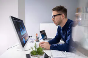 Een man met een bril werkt geconcentreerd achter zijn computer aan data-analyse voor asset management op kantoor. Op het scherm staat: "Analytics", "Audience", "Country", "City", "Status", "Sex", "Age", "Interests", "Collaboration", "Behavior", "Technology", "Mobile", "Custom 1", "Custom 2", "Custom 3", "Income", "Education", "Daily Unique Sales by Country", "Daily Product Sales by Country".