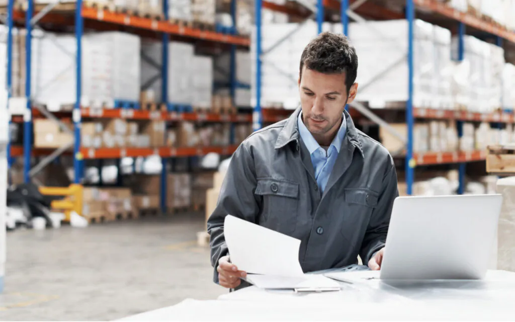 Een magazijnmedewerker in werkkleding beoordeelt documenten bij een laptop ter ondersteuning van SAP FSM in een druk distributiecentrum met hoge stellingen vol voorraadartikelen.