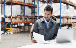 Een magazijnmedewerker bekijkt documenten achter een laptop om het asset management in een distributiecentrum te optimaliseren, omringd door hoge stellingen vol voorraad in een professionele logistieke omgeving.