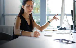 Een zakelijke vrouw neemt aantekeningen terwijl ze naar haar computerscherm wijst in een licht kantoor, wat haar helpt bij efficiënt field service management en digitale werkzaamheden aan haar bureau.
