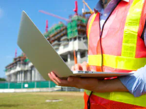 Een technicus in een reflecterend veiligheidsvest bedient een laptop voor **field service management** op een actieve bouwplaats met stellingen en kranen op de achtergrond.
