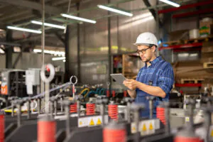 Een technicus met een helm en bril bedient een tablet voor field service management terwijl hij in een industriële fabriekshal met machines en opslagstellingen op de achtergrond staat.
