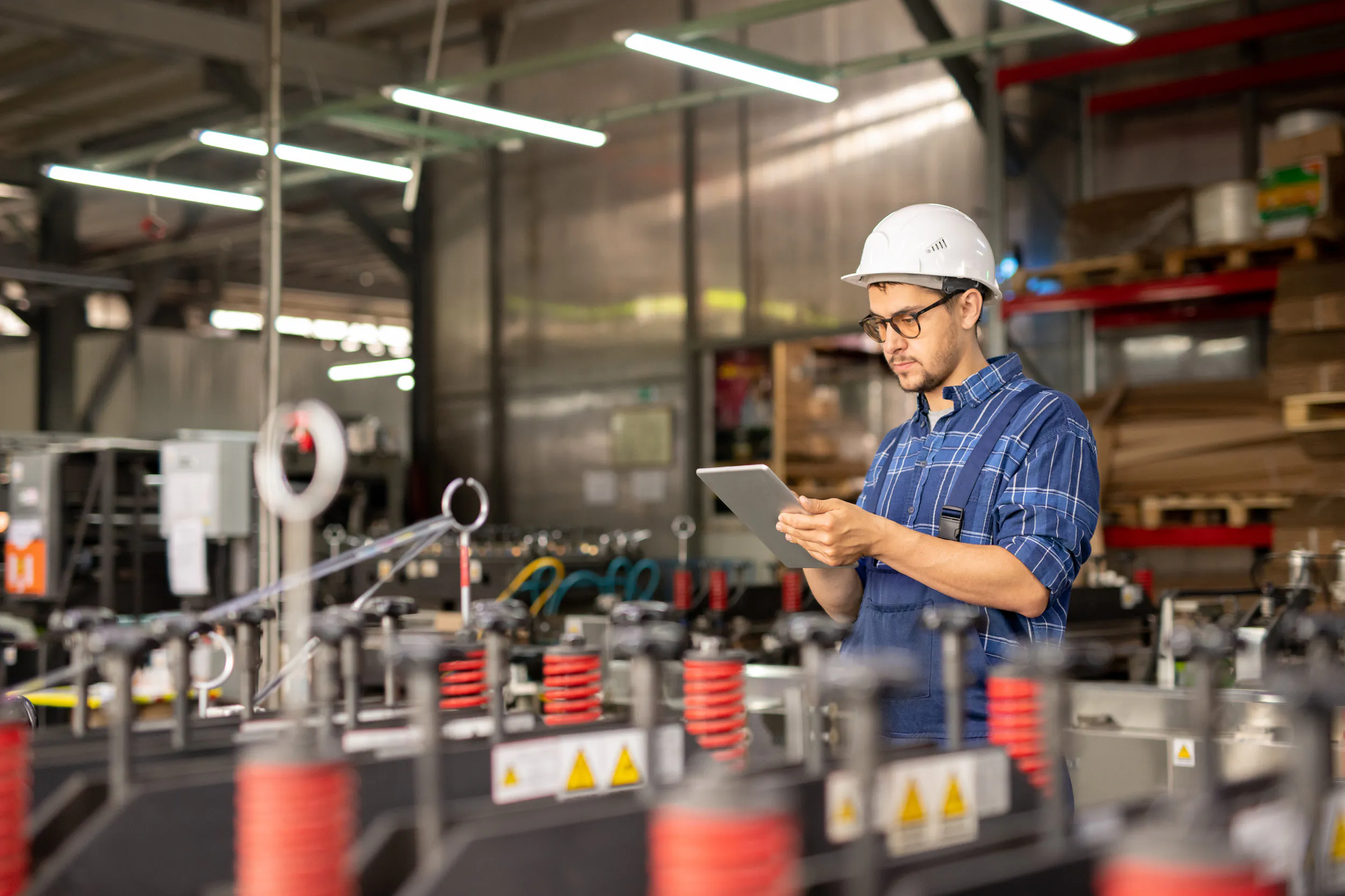 Een technicus met een helm en bril bedient een tablet voor field service management terwijl hij in een industriële fabriekshal met machines en opslagstellingen op de achtergrond staat.