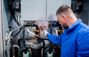 Een technicus in blauwe werkkleding gebruikt een stroomtang voor sap fsm asset management, terwijl hij metingen verricht aan de complexe bedrading en drukmeters van een industriële koelinstallatie. Tekst in de afbeelding: "DANGER" "DANGER" "PRESENZA TENSIONE