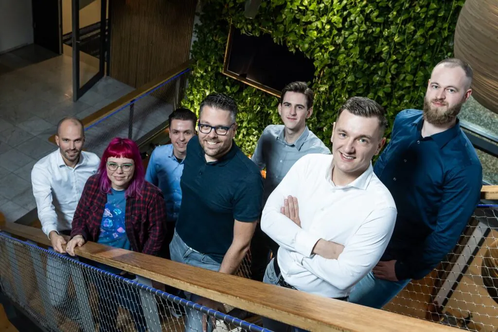 Zes collega's poseren glimlachend op een trap bij een groene muur, wat hun samenwerking binnen **field service management** weerspiegelt in een moderne kantooromgeving.