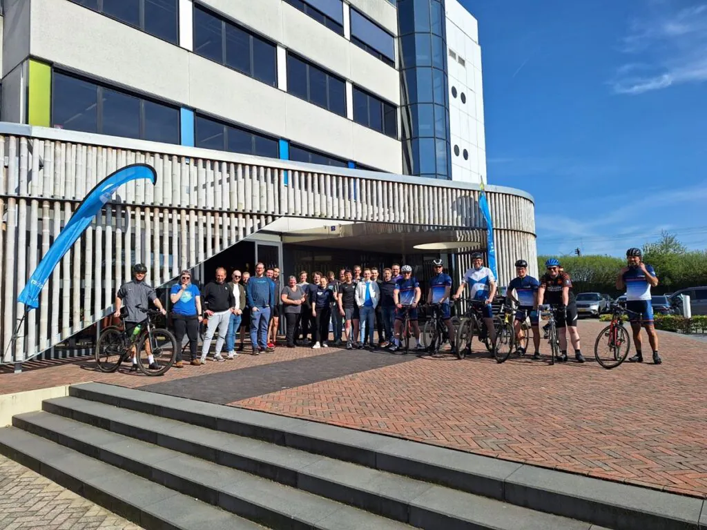 Een groep mensen in wielerkleding en vrijetijdskleding poseert met fietsen voor een modern kantoorpand, passend bij een teamuitje gerelateerd aan SAP FSM en professioneel asset management.