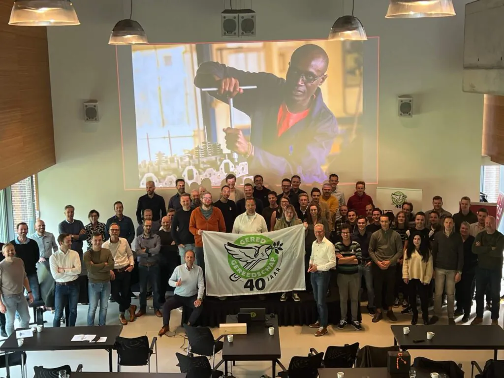 Een grote groep mensen staat in een conferentieruimte en houdt een vlag vast voor een groot scherm met een monteur, wat doet denken aan SAP FSM-oplossingen voor field service management. Tekst op vlag: "GERED GEREEDSCHAP 40 JAAR