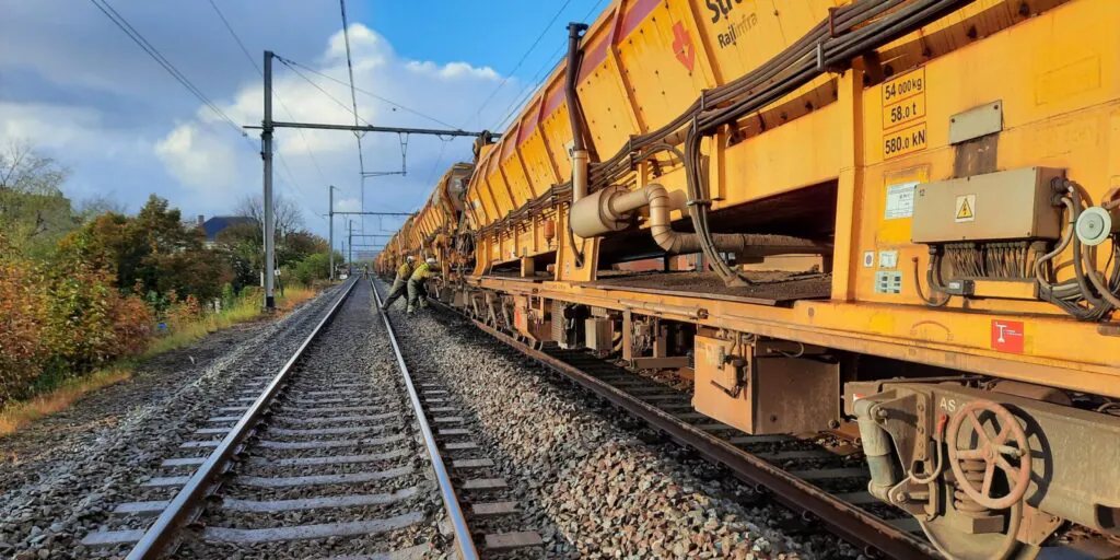 Twee technici voeren onderhoud uit aan een gele werktrein langs het spoor, wat essentieel is voor effectief field service management. Tekst op de trein: "Strukton Railinfra" "54 000 kg" "58.0 t" "580.0 kN" "12" "AS