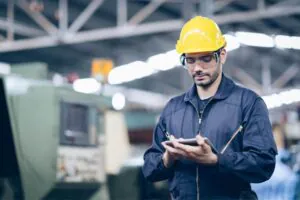 Een technicus met een gele helm en veiligheidsbril gebruikt een tablet voor field service management in een industriële fabriekshal met zware machines op de achtergrond.