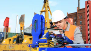 Een technicus met een helm en reflecterend vest gebruikt SAP FSM op een AR-scherm om een kabel te verwijderen op een industriële bouwplaats. Tekst op scherm: "John Doe", "Remove the cable".