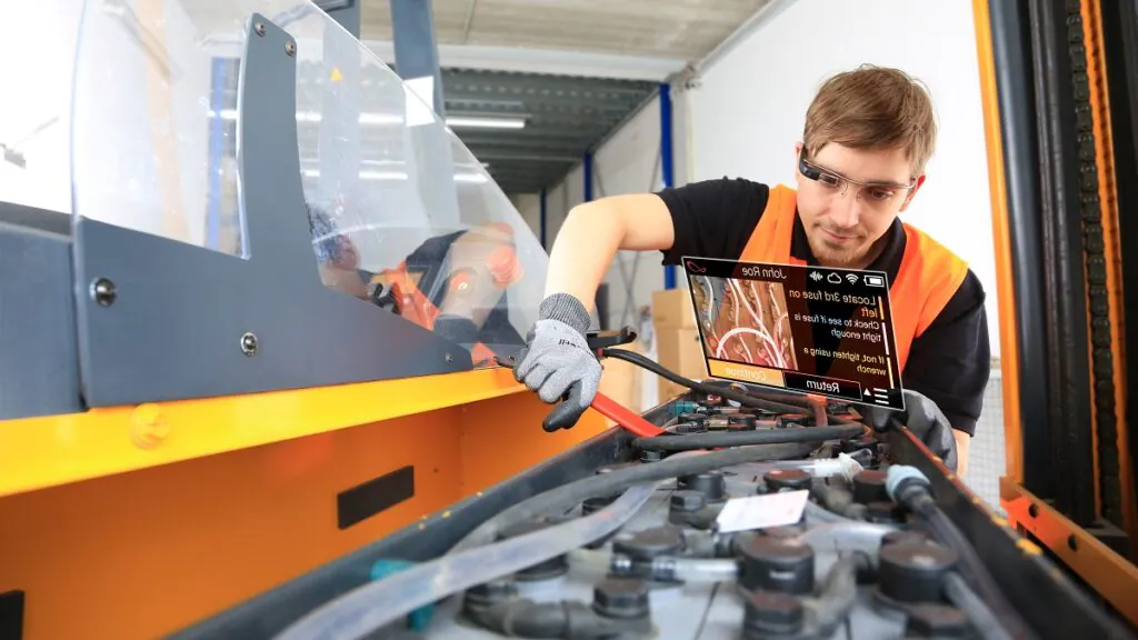 Een monteur met een slimme bril voert onderhoud uit aan een heftruck met behulp van SAP FSM-technologie in een magazijn. Tekst op scherm: "John Roe Locate 3rd fuse on left Check to see if fuse is tight enough If not, tighten using a wrench Continue Return