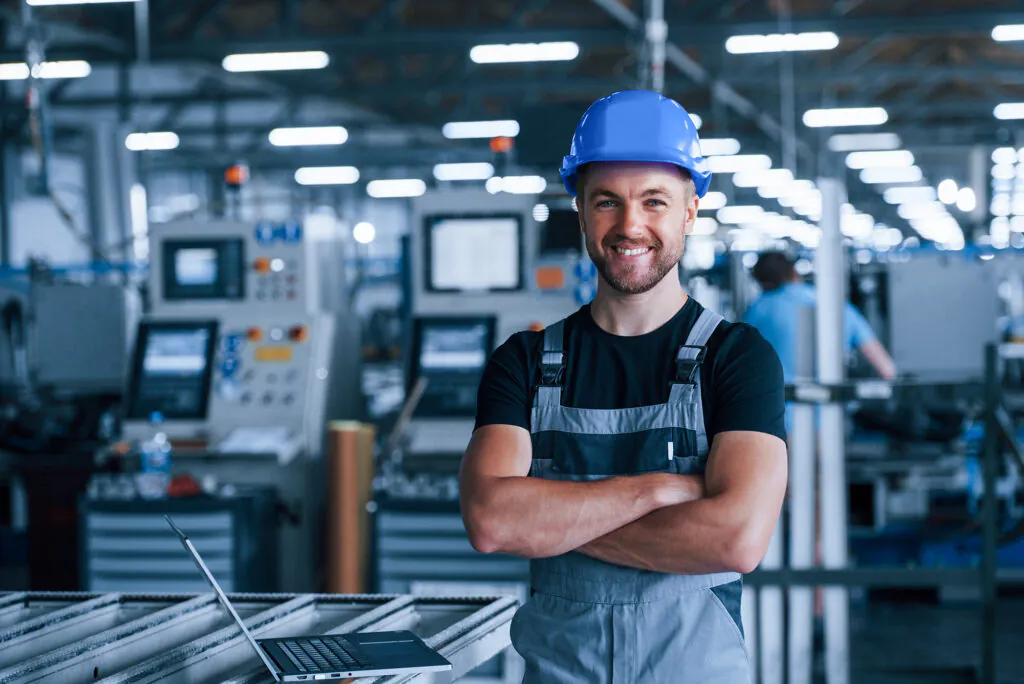 Een glimlachende technicus met een blauwe helm staat met gekruiste armen in een fabriekshal, klaar voor efficiënt field service management bij het onderhouden van de machines op de werkvloer.