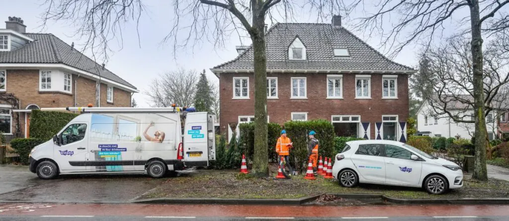 Twee Vitens-monteurs voeren werkzaamheden uit in een woonwijk voor field service management, terwijl ze bij een bedrijfswagen en een personenauto staan voor een bakstenen huis. Tekst op de bus: "je doucht met 70 liter drinkwater dat kan minder!" "water voor nu en later" "Vitens" "V-995-RB" Tekst op de auto: "water voor nu en later" "Vitens