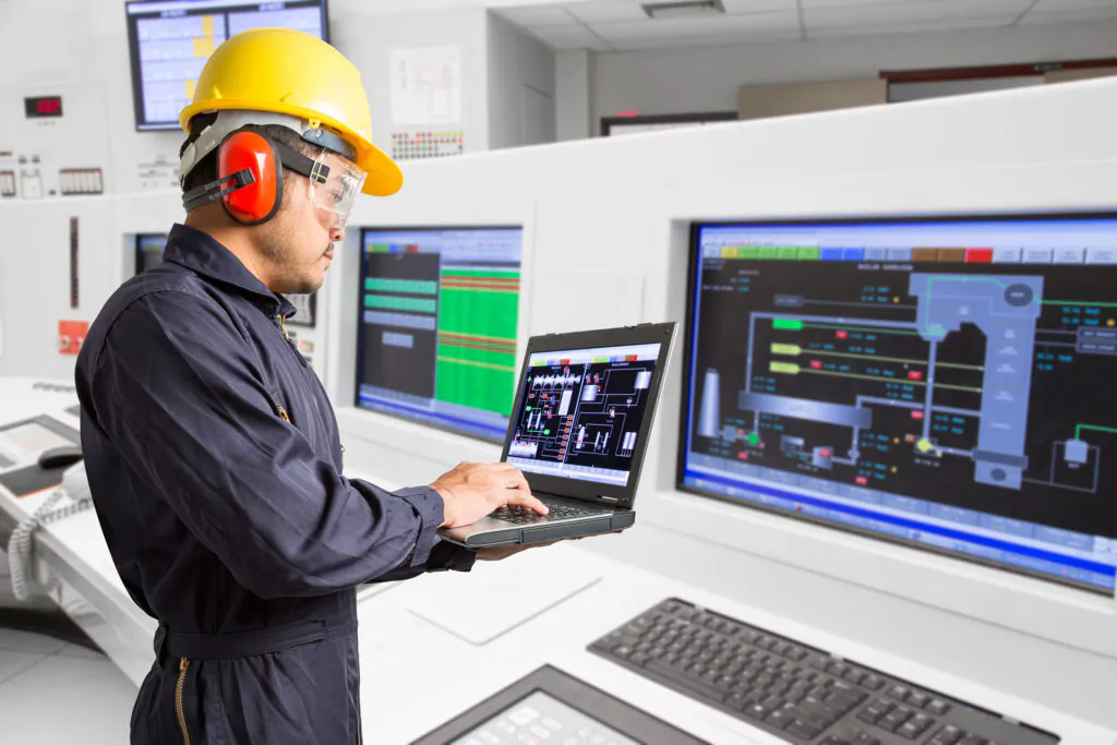 Een technicus met helm en gehoorbescherming gebruikt een laptop voor asset management in een centrale controlekamer vol met computerschermen die industriële procesgegevens en schema's weergeven.