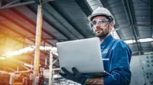 Een technicus in een blauwe overall met helm en veiligheidsbril houdt een laptop vast om SAP FSM-gegevens te controleren in een grote, industriële fabriekshal met een hoog plafond.